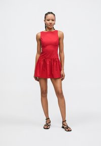 Červené šaty bez rukávů s přiléhavým živůtkem a nabíranou sukní, vybavené nastavitelnými šňůrkami v pase. Hladká látka s matným povrchem.