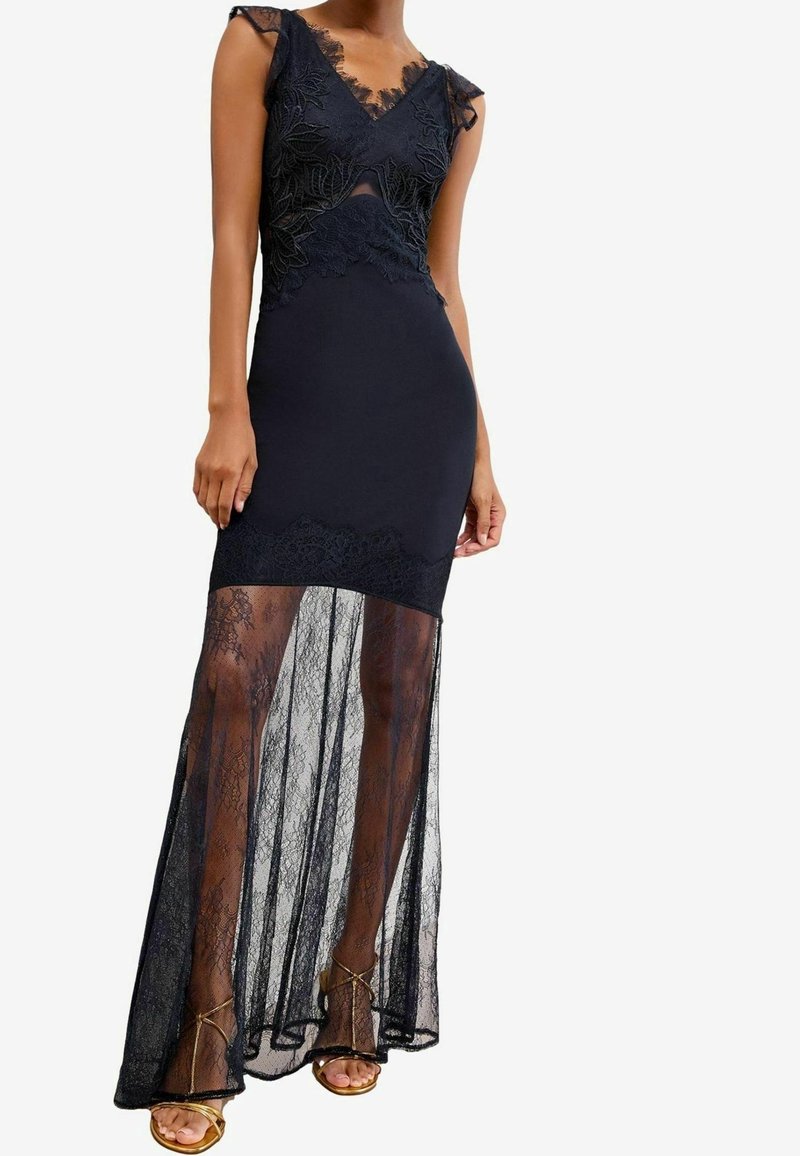 Femme portant une robe noire en dentelle sans manches avec un corsage ajusté et une superposition de jupe en dentelle transparente, associée à des talons ouverts dorés.
