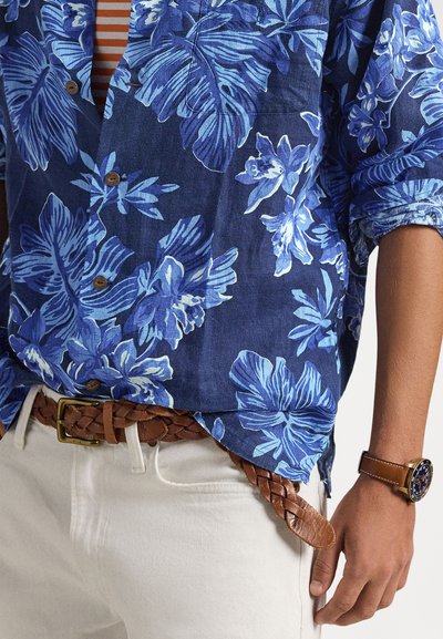 Μπλε πουκάμισο με λουλουδάτο σχέδιο πάνω από καφέ ριγέ τοπ, συνδυασμένο με λευκό παντελόνι, καφέ πλεκτή ζώνη και καφέ δερμάτινο ρολόι στον αριστερό καρπό.