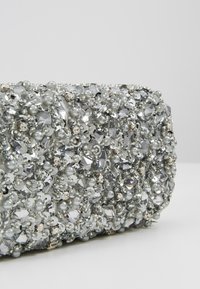 Pochette perlée recouverte de diverses ornements argentés et perleux, présentant un mélange de textures lisses et facettées.