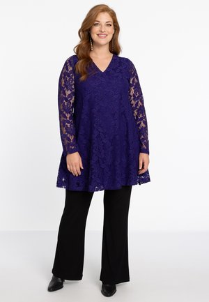 Lila Spitze-Tunika mit langen, transparenten Ärmeln, V-Ausschnitt, ausgestelltem Saum, kombiniert mit schwarzen Hosen und schwarzen, hohen Schuhen. Verzierte Details mit aufwendigem Blumenmuster.