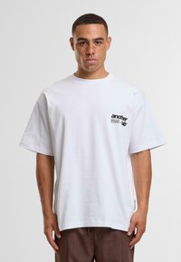 Tricou alb din bumbac cu o croială lejeră, mâneci scurte și un mic logo grafic negru pe partea stângă a pieptului. Textură moale, decolteu tip crew.