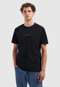 Jonge man met krullend haar, gekleed in een zwart T-shirt met de tekst "PURE PATH" en blauwe spijkerbroek, staande met één hand in zijn zak tegen een effen achtergrond.