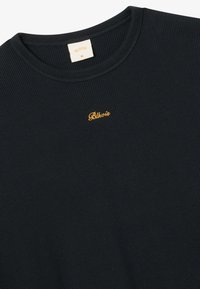 Maglietta a maniche lunghe in maglia waffle nera con scollatura rotonda e piccolo logo giallo "Blkvis" ricamato sul petto. Etichetta di taglia XL all'interno del colletto.