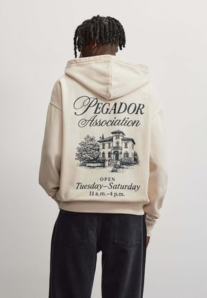 Person seen from behind wearing a beige hoodie with "Pegador Association" text and a house illustration on the back.