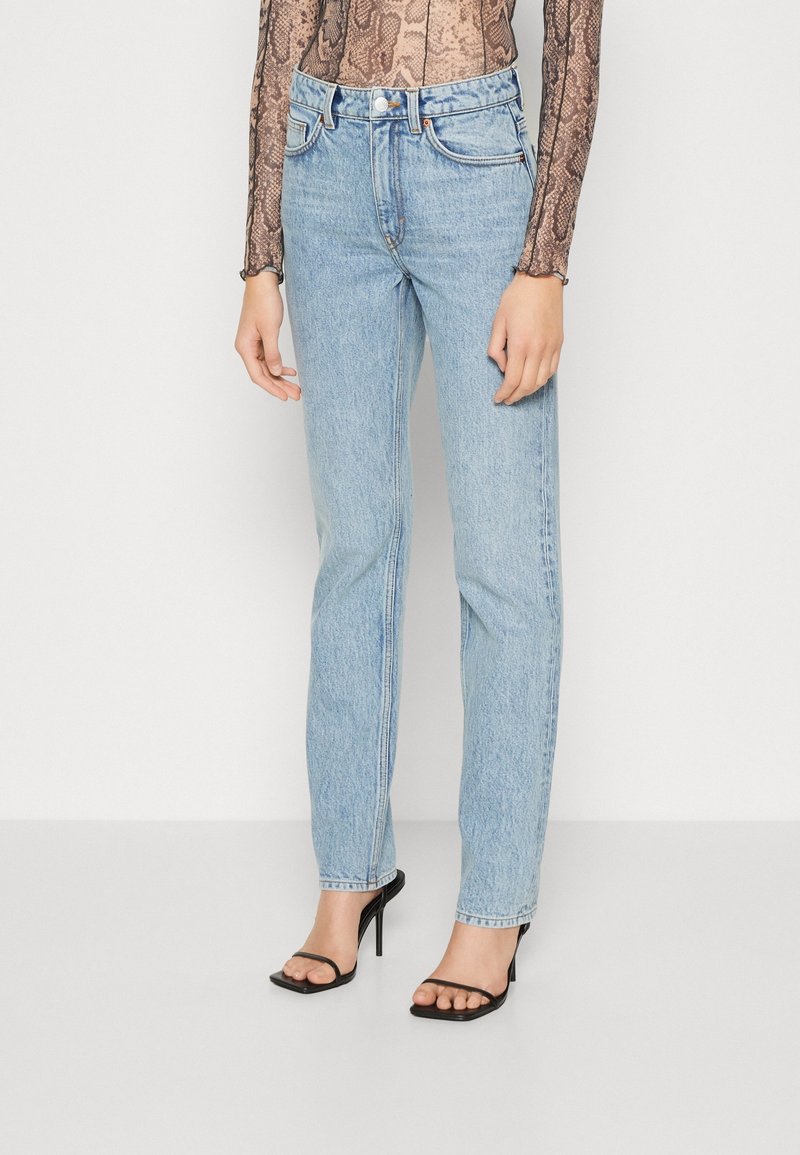 Jean bleu clair taille haute à jambe droite, porté avec des sandales noires à talons hauts et bouts ouverts, ainsi qu'un top à manches longues imprimé serpent sur fond blanc.