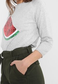 Maglietta grigia a maniche lunghe con un design a forma di fetta di anguria decorata in paillettes rosse e verdi sul davanti, abbinata a pantaloni alti di colore verde scuro.