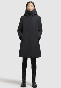 Manteau long noir avec un col haut, fermeture éclair à l'avant et poches latérales. Il présente un design élégant et est fabriqué dans un tissu lisse et durable.