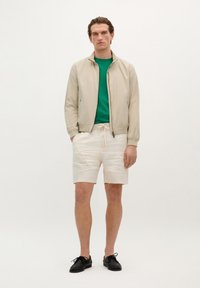 Chaqueta bomber beige, camiseta verde, pantalones cortos crema y zapatos negros. Tejido suave, corte relajado, cintura con cordón, minimalismo en los detalles.