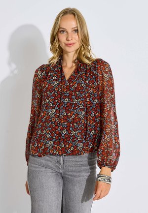 Mujer con una blusa roja de manga larga con estampado floral y jeans grises, con cabello rubio ondulado y pulseras plateadas en su muñeca derecha.