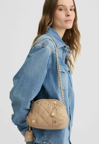 Beige gesteppte Handtasche mit goldfarbenen Beschlägen, Kettenriemen und dekorativen Knöpfen. Die glatte Textur und die abgerundete Form verleihen ihrem Design Flair.