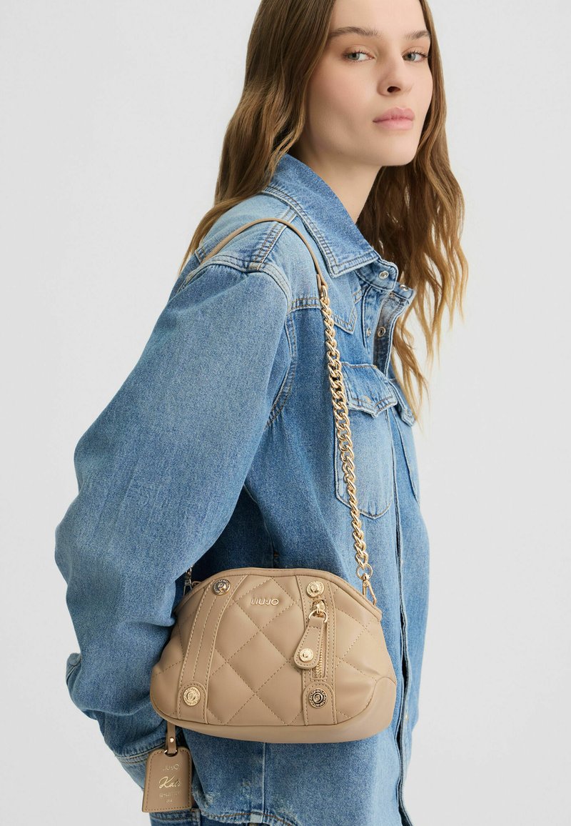 Beige gesteppte Handtasche mit goldfarbenen Beschlägen, Kettenriemen und dekorativen Knöpfen. Die glatte Textur und die abgerundete Form verleihen ihrem Design Flair.