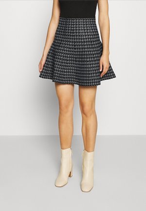 Woman wearing a black and gray houndstooth patterned skirt, black top, and cream ankle boots standing against a plain background.