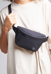 Sac bandoulière noir en tissu texturé, doté d'une fermeture éclair et d'un petit logo. La sangle ajustable permet de le maintenir autour du corps.