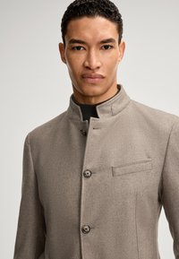 Hellbraune Jacke aus Wollmischung mit hohem Kragen, zwei Knöpfen und einer Brusttasche. Der Stoff hat eine glatte Textur und eine taillierte Passform.