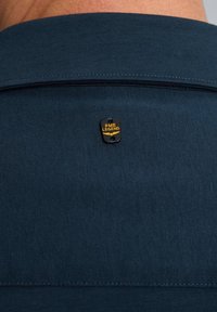 Tamno plava tkanina s teksturiranim završetkom, s crnom etiketom na kojoj piše "PME LEGEND" u žutoj boji. Kragnasti dizajn s vidljivim detaljima šivanja.