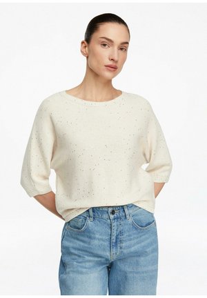 Kobieta z ciemnymi włosami związanymi do tyłu, ubrana w kremowy sweter dziany z krępanym wzorem z rękawami do łokci i niebieskie, wysokowalne dżinsy, stojąca.