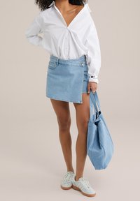 Falda envolvente de mezclilla con un lavado claro, bolsillo lateral y detalles de hardware plateado, combinada con una camisa blanca y un bolso tote azul claro.
