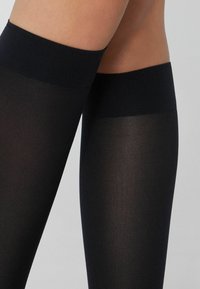 Černé punčochy do stehna s hladkým, průhledným povrchem a širokým, pevným černým páskem na vrcholu. Žádné viditelné vzory ani kovové doplňky.