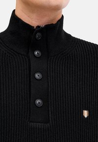 Schwarzer gerippter Strickpullover mit hohem Kragen, ausgestattet mit vier schwarzen Knöpfen und einem kleinen, kontrastierenden Logo auf der Brust.