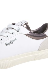 Baskets basses blanches avec lacets blancs, détail talon gris foncé métallisé, semelle beige et logo métallique "Pepe Jeans" sur le côté.