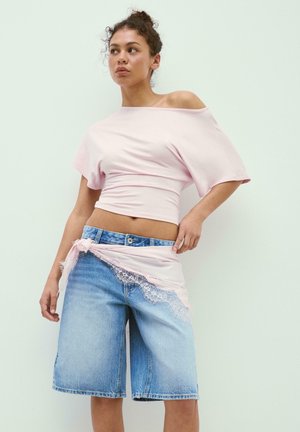 Femme portant un haut rose clair à épaules dénudées et un short en jean longueur genou avec un foulard en dentelle rose noué autour de la taille.