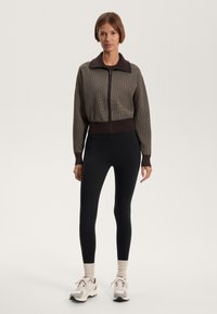 Brown ribbed zip-up jacket with a black collar, paired with black leggings and beige sneakers. The outfit features a fitted design and contrasting colors.