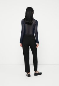 Femme aux longs cheveux noirs portant un haut rayé transparent bleu marine, un pantalon noir ajusté et des mocassins noirs, se tenant de dos sur un fond blanc.