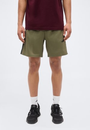 Hombre con pantalones cortos deportivos verde oliva, camiseta granate, calcetines blancos de Adidas y zapatillas negras para correr, de pie frente a un fondo liso.