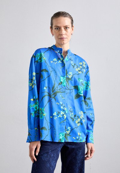Blauw bloemetjes overhemd met groene en gele bloemenpatronen, lange mouwen en een mandaryn collar. Draagt met donkere blauwe broek.