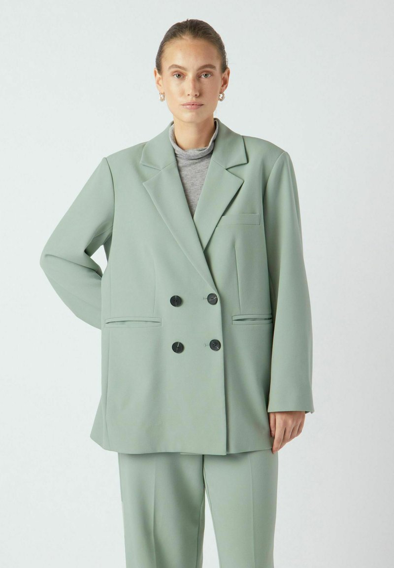Dobbelspent blazer i mykt mintgrønt stoff med to lommer foran, sorte knapper og en avslappet passform. Halsrør under i grått.