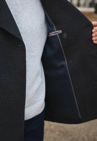 Person holding open a dark checkered coat to reveal a navy blue interior lining and a buttoned inner pocket, wearing a light gray sweater underneath.