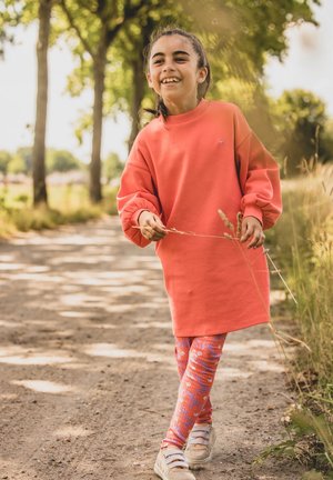 Koraal oversized sweatshirt, patroonlegging in oranje en roze, en beige sneakers. Gevestigd op een zandpad omzoomd met bomen.
