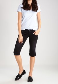 Ljusblå V-hals t-shirt i kombination med svarta capri-jeans. Modell som bär svarta slip-on skor och står på en neutral bakgrund.