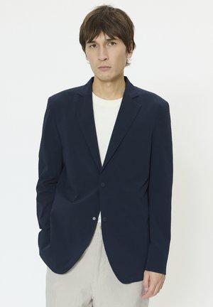Sötétkék blézer texturált anyagból, egygombos záródással és bevágott hajtókákkal, világos színű inggel és csíkos nadrággal párosítva.