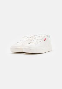 Kaksikkaat valkoiset matalavartiset lenkkarit valkoisilla nauhoilla, vihreällä brändäyksellä kielekkeessä ja pienellä punaisella tarralla sivussa valkoista taustaa vasten.