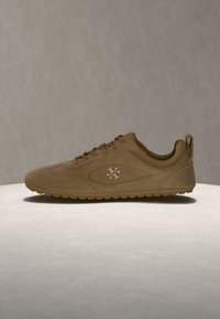 Baskets en daim marron avec lacets et logo de marque sur le côté, placées sur une surface claire sur un fond neutre uni.