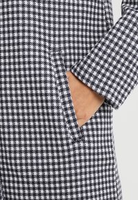 Ruka částečně v kapse rukávu černo-bílého kabátu s kostkovaným vzorem houndstooth a viditelný boční detail.
