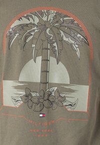 Olivenfarget t-skjorte med et palmetre-motiv, stiliserte blomster og en solnedgang. Teksten inkluderer "HILFIGER NEW YORK 1985" med logo-detajer.