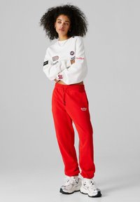 Γυναίκα με κατσαρά μαλλιά στέκεται με σταυρωμένα τα χέρια, φορώντας λευκό φούτερ με λογότυπα, κόκκινη φόρμα RedBull Racing και λευκά αθλητικά παπούτσια.