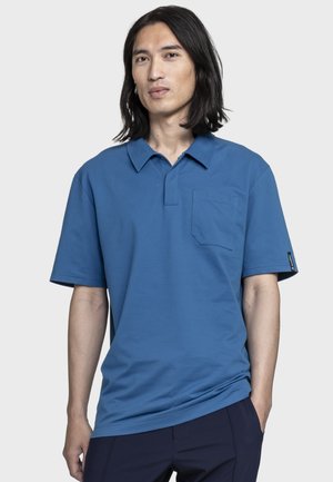 Junger Mann mit langen schwarzen Haaren, der ein blaues Polohemd mit kurzen Ärmeln und Brusttasche sowie dunkle Hosen trägt, vor einem einfachen Hintergrund.