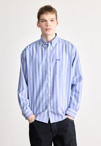 Gestreiftes blaues Hemd aus Stoff mit langen Ärmeln, einer Tasche auf der linken Brust und einem kleinen Logo-Tag. Dunkle Jeans darunter.