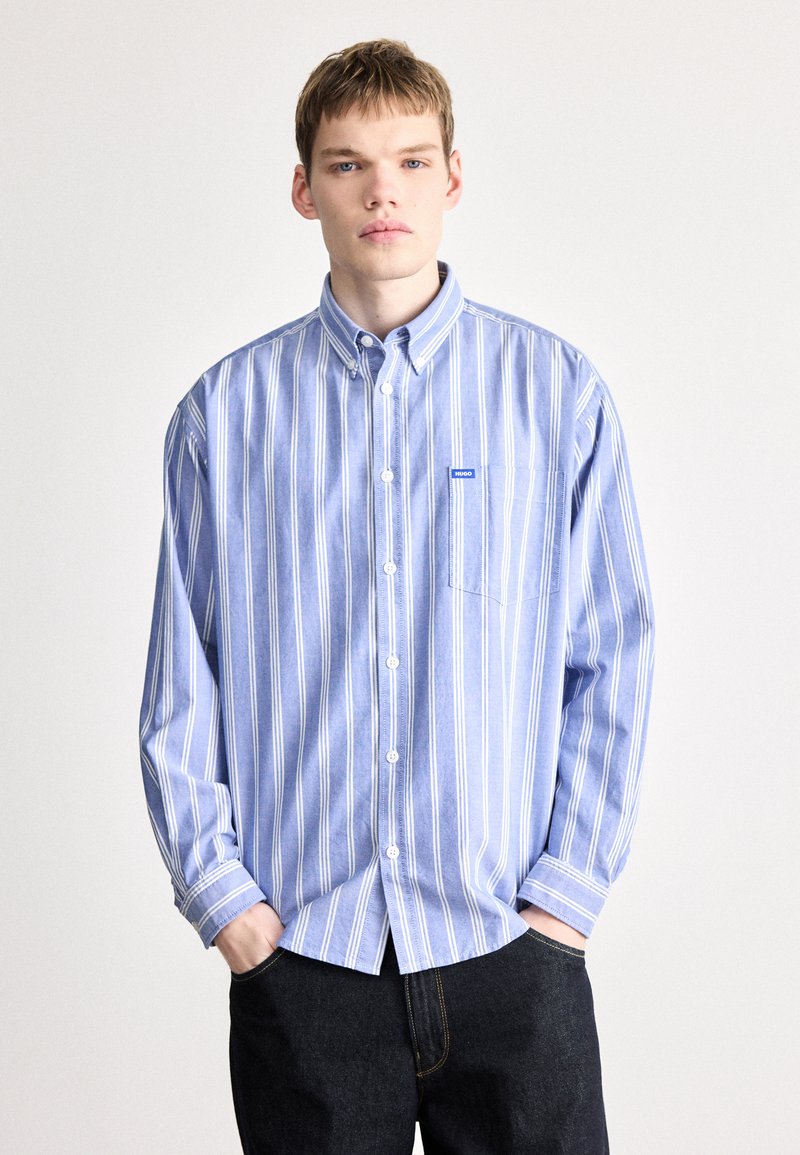 Gestreiftes blaues Hemd aus Stoff mit langen Ärmeln, einer Tasche auf der linken Brust und einem kleinen Logo-Tag. Dunkle Jeans darunter.