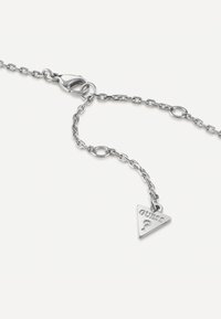 Bracelet en chaîne argentée avec des maillons interconnectés, un fermoir en forme de homard et une étiquette triangulaire avec "GUESS ?" embossé dessus.