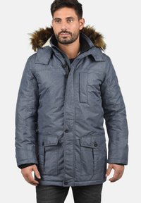 Blå isoleret parkacoat med hætte i faux fur, forlommer, knaplukning og en tekstureret overflade. Designet til varme og komfort.