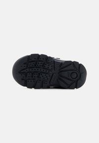 Semelle de chaussure en caoutchouc noir avec un motif de semelle texturé, comportant des rainures et des accents circulaires pour la prise et la traction. Étiquette indiquant la marque présente.