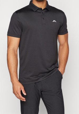 Mężczyzna w czarnej koszulce polo z krótkim rękawem, trzema guzikami i białym logo na lewej piersi, w czarnych spodniach, z ręką w kieszeni.