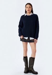 Maglione navy a treccia, camicia bianca sotto, gonna mini a quadretti e stivali marroni al ginocchio con fibbie e tessuto texture.