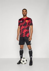 Rood-zwart patroon voetbalshirt met korte mouwen, gecombineerd met zwarte korte broek en sokken. Een voetbal ligt bij de voeten.