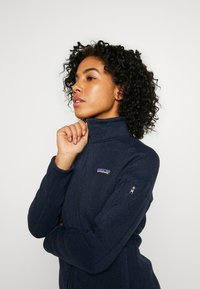 Giacca in pile con zip di colore blu navy, con superficie testurizzata, colletto alto e logo sul petto. Presenta una tasca con zip sul maniche sinistro.